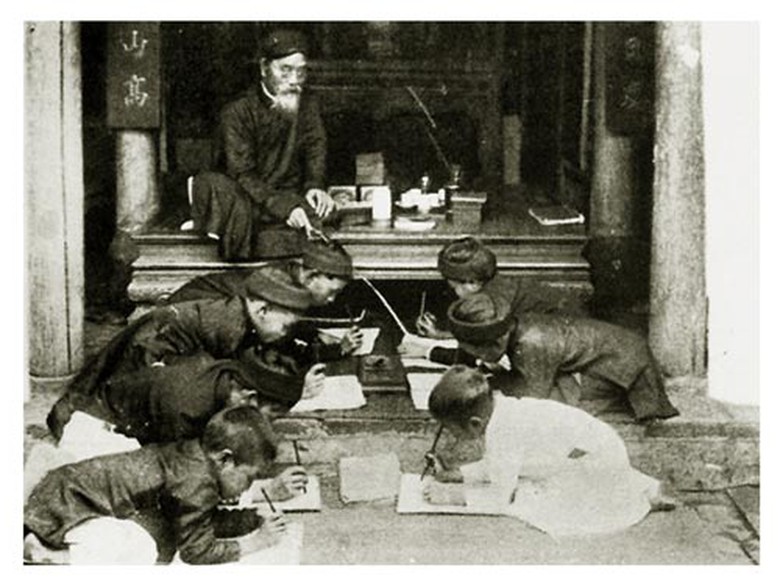 Thầy cầm roi, ngồi tr&ecirc;n sạp gụ chỉ dẫn học tr&ograve; viết chữ Nho. Theo Nho gi&aacute;o ng&agrave;y xưa, học sinh phải k&iacute;nh trọng thầy gi&aacute;o hơn cha mẹ, thầy chỉ sau vua, theo thứ bậc 