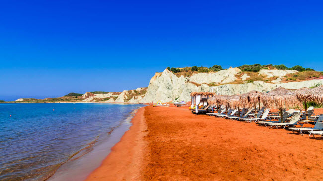 B&atilde;i biển Xi, Hi Lạp: B&atilde;i biển nằm tr&ecirc;n h&ograve;n đảo Kefalonia nổi tiếng với c&aacute;c m&agrave;u cam sặc sỡ. Nước biển ở đ&acirc;y ấm, n&ocirc;ng v&agrave; s&oacute;ng nhẹ. M&agrave;u hồng của c&aacute;t tương phản với v&aacute;ch đ&aacute; m&agrave;u trắng, tạo n&ecirc;n khung cảnh tuyệt mỹ.
