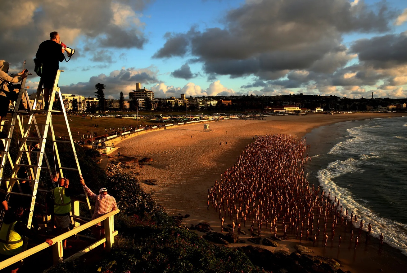 Quy định cho ph&eacute;p khỏa th&acirc;n tr&ecirc;n b&atilde;i biển Bondi chỉ c&oacute; hiệu lực đến 10 giờ, n&ecirc;n nếu kh&ocirc;ng muốn bị phạt nh&oacute;m người tham gia chụp ảnh phải đi sớm v&agrave; thật khẩn trương.
Từ 4 giờ s&aacute;ng, &ocirc;ng Tunick đ&atilde; đứng tr&ecirc;n cầu thang&nbsp;m&agrave;u da cam, chỉ đạo người mẫu di chuyển qua loa. Trong l&uacute;c tập xếp đội h&igrave;nh, một số người mẫu quấn tạm khăn tắm quanh người cho ấm, một kh&aacute;c tranh thủ chụp v&agrave;i tấm ảnh l&uacute;c mặt trời l&ecirc;n.