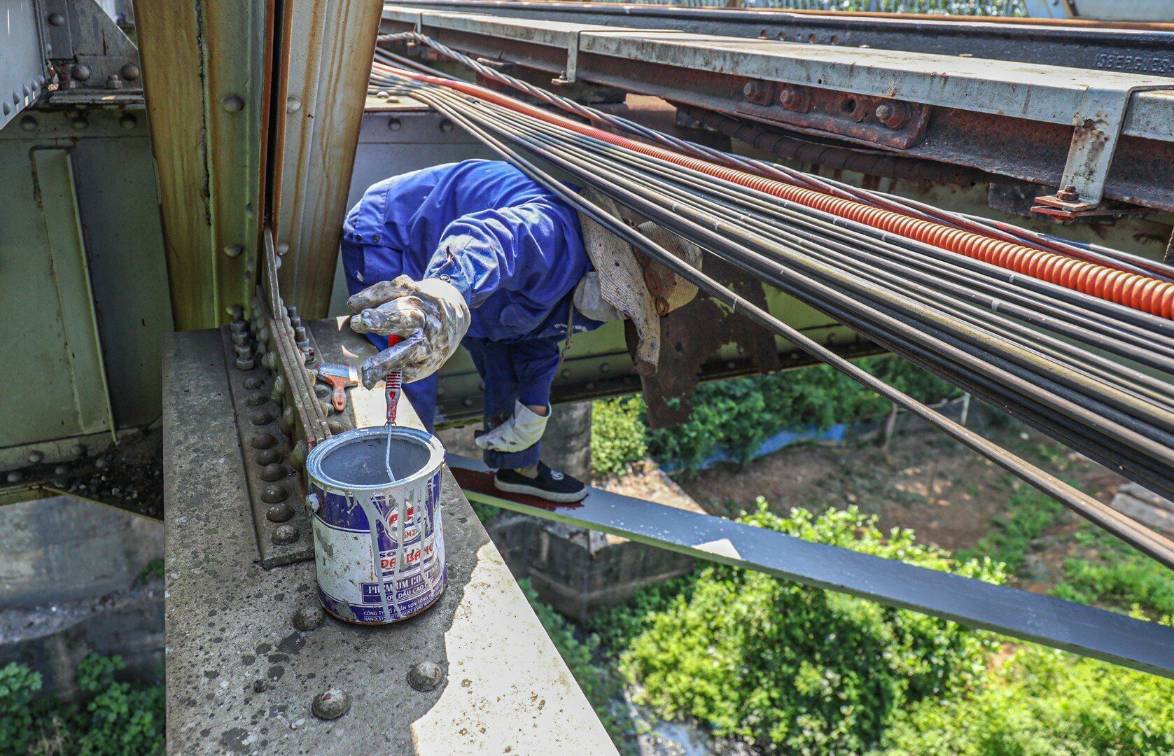 Trong nắng gắt, c&aacute;c thanh th&eacute;p tr&ecirc;n cầu c&agrave;ng th&ecirc;m n&oacute;ng r&atilde;y, hơi n&oacute;ng phả ra hầm hập. Dẫu vậy, c&aacute;c c&ocirc;ng nh&acirc;n vẫn đeo, b&aacute;m c&aacute;c thanh th&eacute;p, sơn cẩn thận, sơn phủ cả những vị tr&iacute; &ldquo;hiểm h&oacute;c&rdquo;, kh&oacute; nh&igrave;n thấy từ ngo&agrave;i đường bộ v&agrave;o, hay từ tr&ecirc;n đường sắt xuống. Ảnh: Nữ c&ocirc;ng nh&acirc;n đứng cheo leo tr&ecirc;n c&aacute;c thanh th&eacute;p nhỏ, sơn ph&iacute;a dưới đường sắt